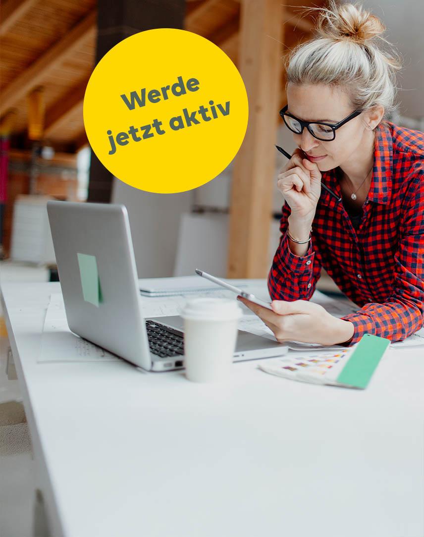 Frau am Schreibtisch vor Laptop schaut aufs Smartphone