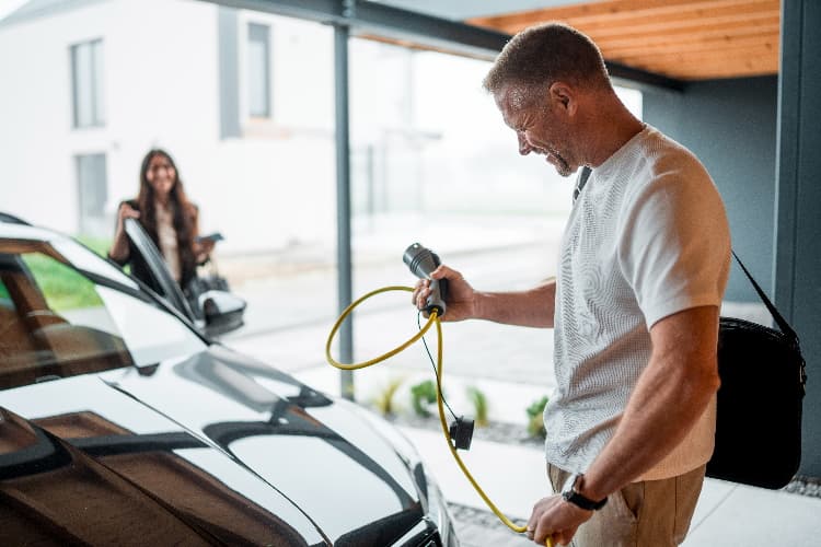 Mann schließt ein Elektroauto an einer Wallbox an. Im Hintergrund steigt eine Frau aus dem Auto.