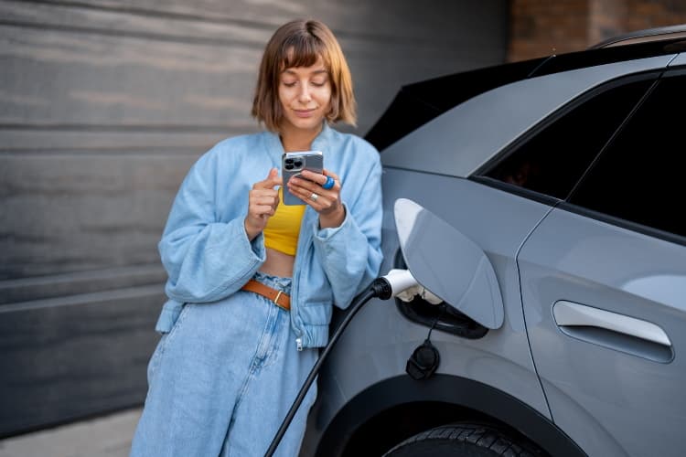 Frau bedient Handy angelehnt an einem E-Auto.