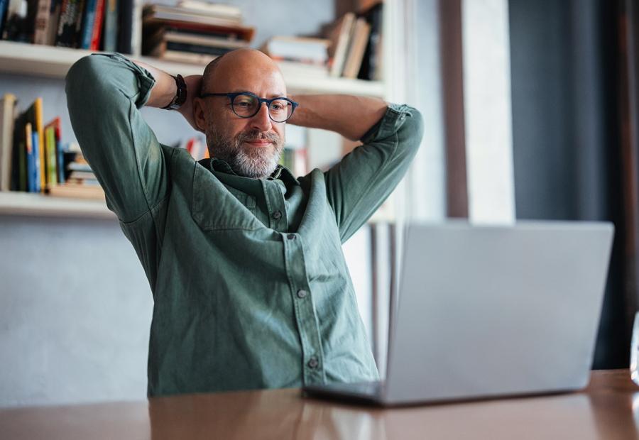 Mann schaut in seinen Laptop und verschränkt zufrieden seine Hände hinter dem Kopf