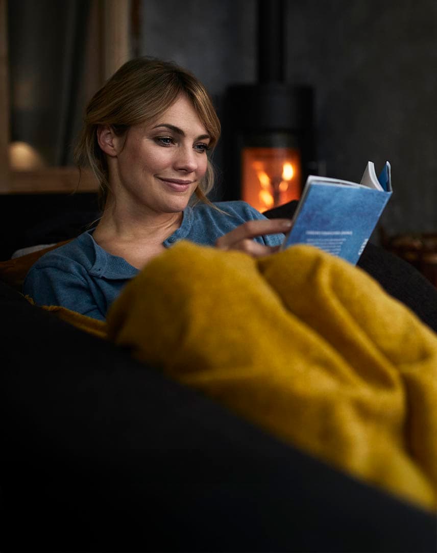 Eine Frau sitzt entspannt auf dem Sofa und liest, im Hintergrund brennt der Kamin