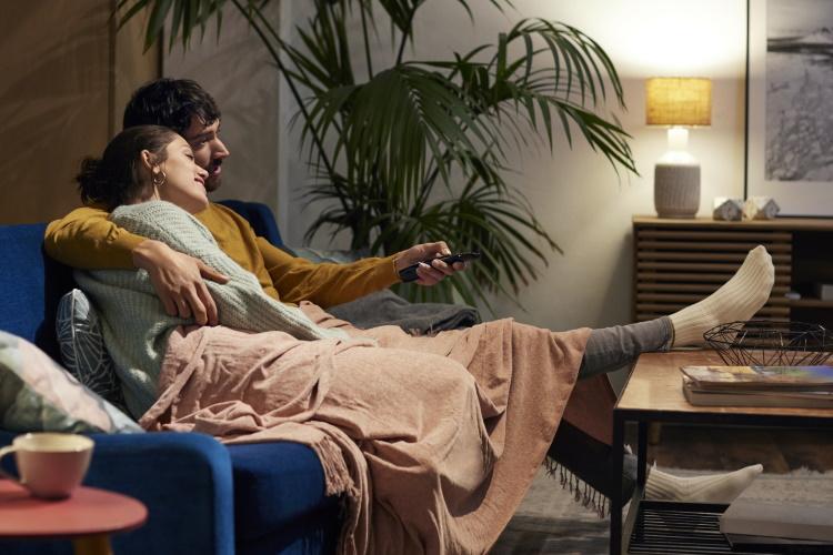 Pärchen sitzt gemütlich auf der Couch und schaltet den Fernseher mit einer Fernbedienung an