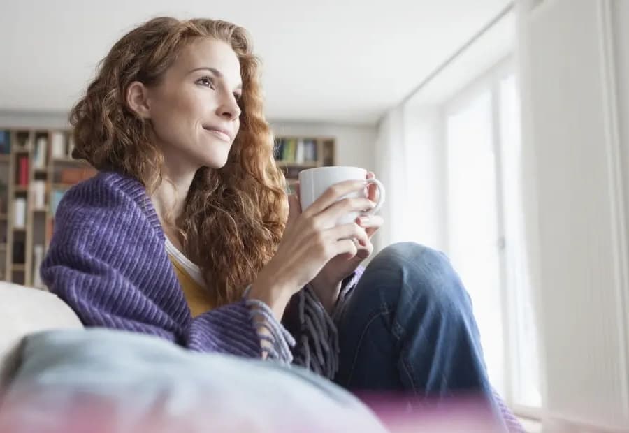 Frau sitzt mit zufriedenem Gesichtsausdruck auf der Couch und trinkt in Ruhe Kaffee