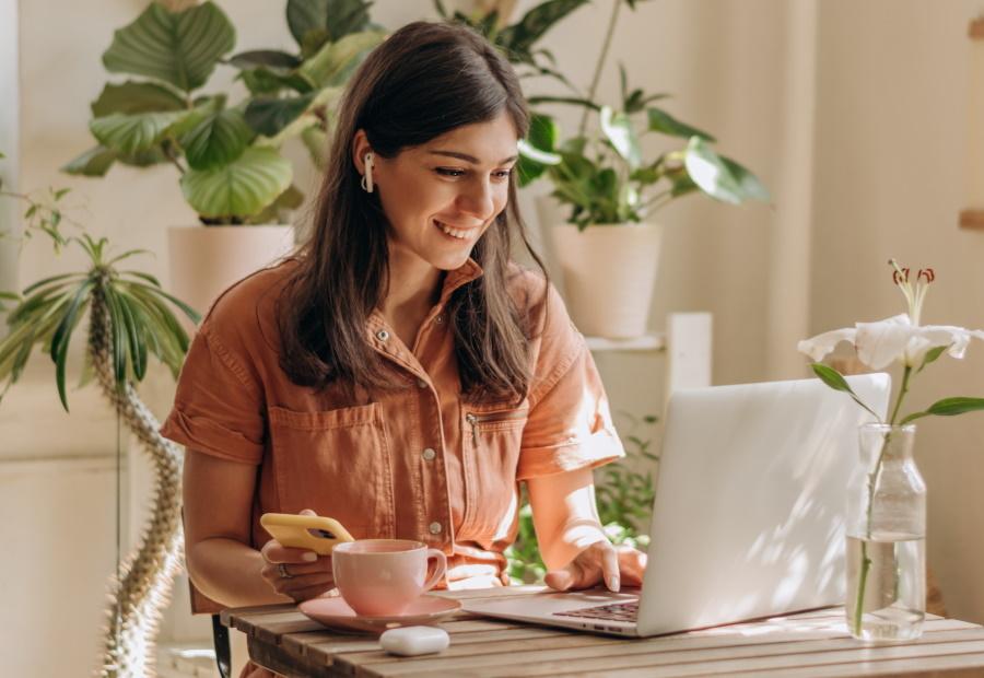 Junge Frau schaut lächelnd auf ihren Laptop bzw. ihr Handy, während sie mit Kopfhörern im Ohr telefoniert