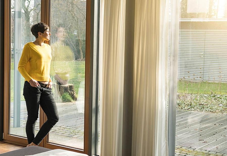 Frau steht am Fenster und schaut in ihren sonnendurchfluteten Garten