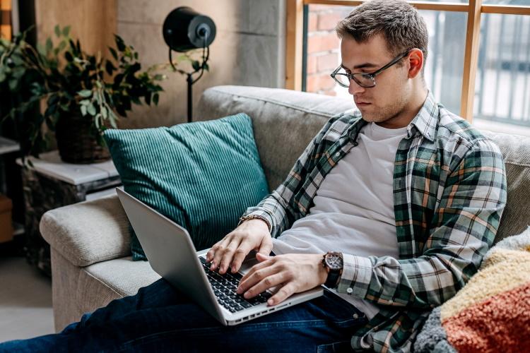 Junger Mann sitzt auf der Couch beim Fernstudium an seinem Laptop