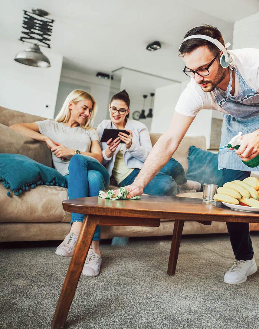 Zwei Freundinnen schauen auf ein Tablet, ein Mann putzt derweil den Couchtisch